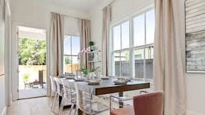 A dining area with a long table set for six, clear chairs, and a pink chair. There are curtains on the windows, and a plant on the table.