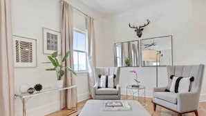 A modern living room with a grey sofa, two armchairs, a glass coffee table, and a large mirror. There is a decorative antler on the wall and a plant on the windowsill.