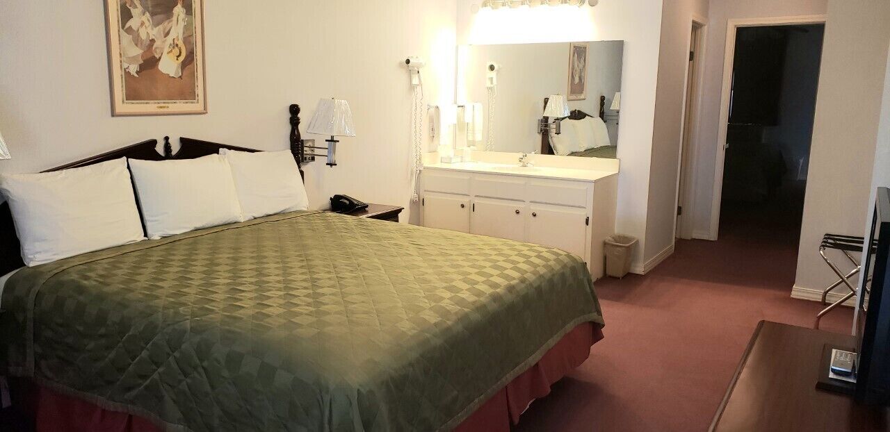 A hotel room with a bed, a bedside table, a mirror, a vanity with a sink, and a door leading to another room.