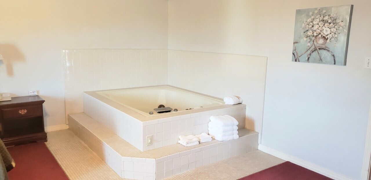 A bathroom with a freestanding bathtub, white tiled walls, a wooden nightstand, and a painting on the wall.