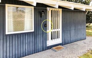 Een klein, laag gebouw met een donkere buitenkant, een raam met een witte omlijsting, een deur met verticale spijlen en een klein portaal.