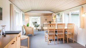 A cozy room with a wooden ceiling, a dining table with chairs, a wicker chair, and a small side table with a candle. - Secluded Pine Tree Cottage-by Traum (Blavand)