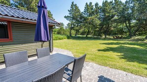 A patio with a table and chairs, an umbrella, and a wooden cabin in the background. - Secluded Pine Tree Cottage-by Traum (Blavand)