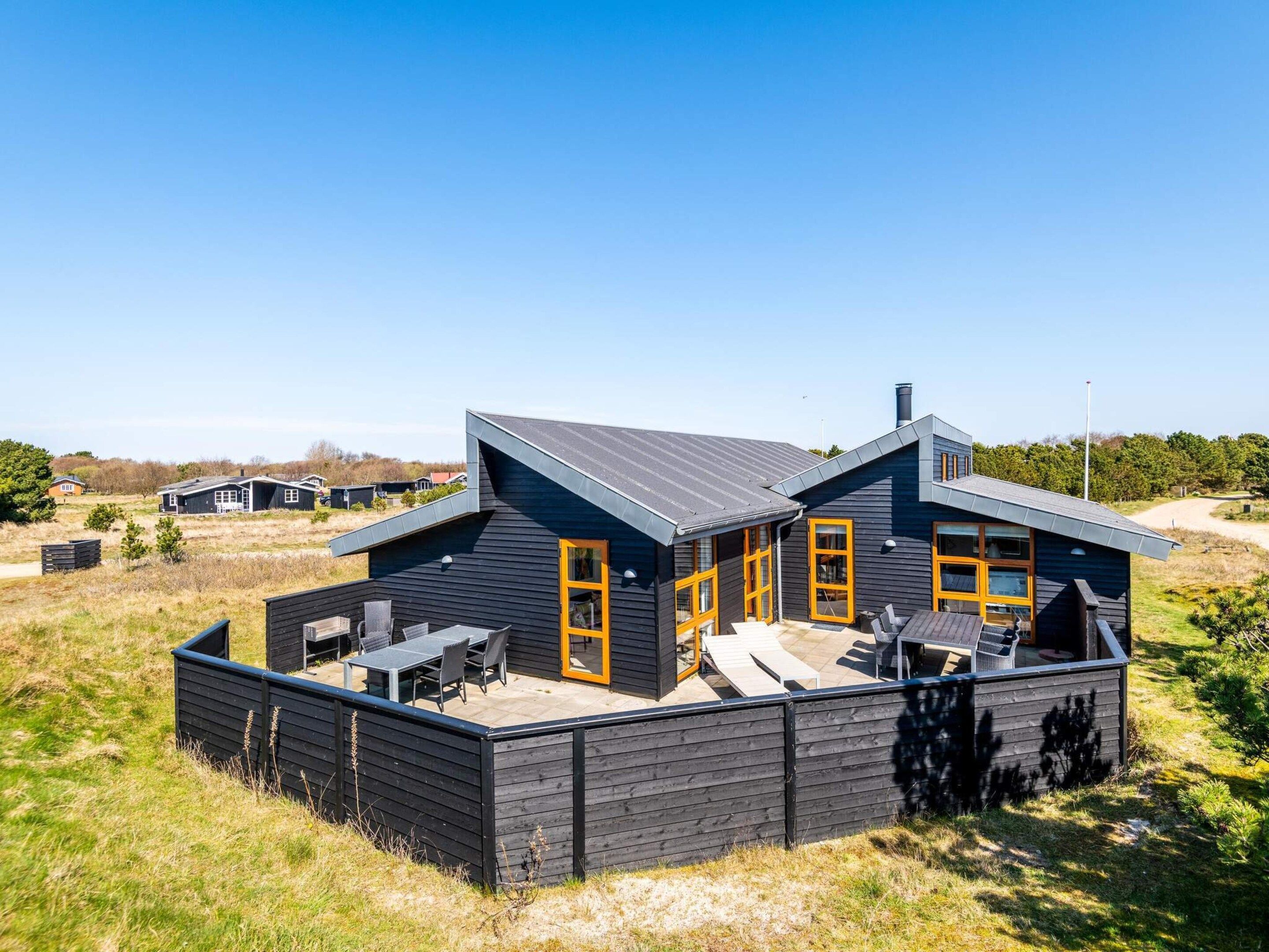 A modern two-story house with a flat roof, large windows, and a patio area with outdoor furniture.