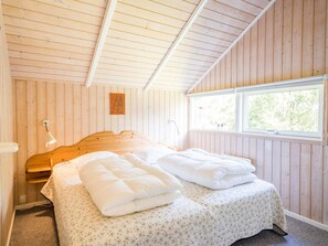 A bedroom with a wooden sloped ceiling, a bed with white bedding, a nightstand, a lamp, and a window with a view of trees.