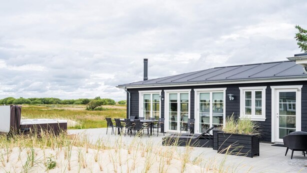 A black building with large windows and a chimney, surrounded by sand dunes and vegetation. - Holiday Home in Blåvand (Blavand)