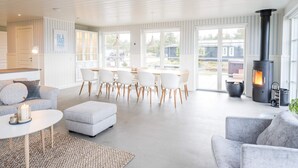 A modern living room with a fireplace, a dining area with white chairs, and a large window providing a view of the outside. - Holiday Home in Blåvand (Blavand)