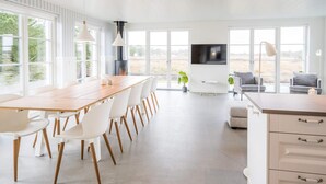 A spacious living room with a long wooden dining table, white chairs, a large TV, and a fireplace. - Holiday Home in Blåvand (Blavand)