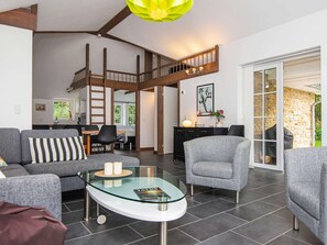 Un vaste espace de vie doté d’un plafond haut, une chambre en mezzanine, un coin repas et une vue sur un mur de pierre et la verdure environnante.