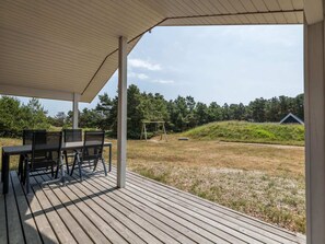 An outdoor patio with a table and chairs, a swing set, and a view of a grassy area with trees and a building in the background.