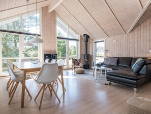 Ein geräumiger Wohnbereich mit Holzbalkendecke, großen Fenstern, einem Kamin, einem Esstisch mit Stühlen und einem schwarzen Ledersofa.