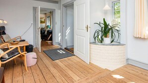 Una habitación con suelo de madera, una puerta blanca, una mesita con una silla, una planta en maceta y una ventana con cortinas.