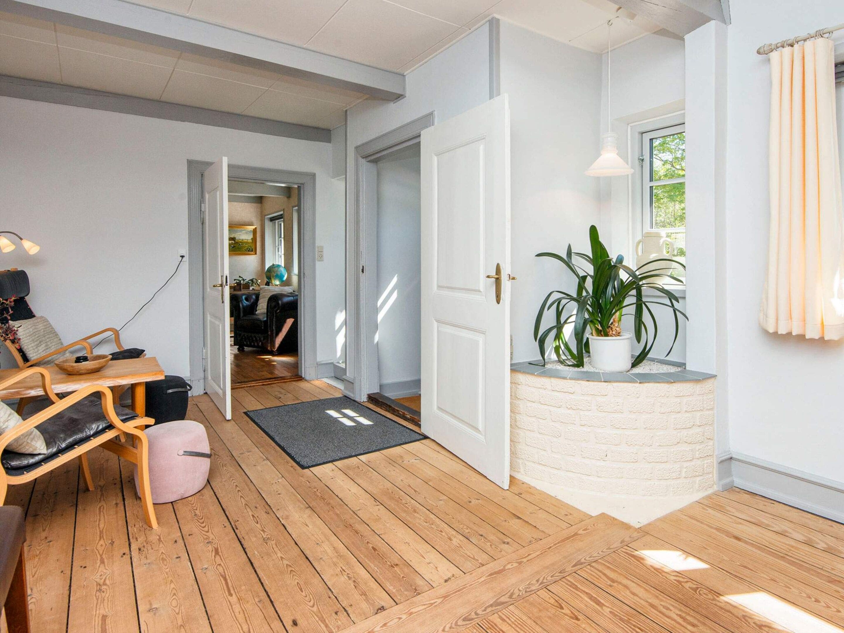Una habitación con piso de madera, una puerta blanca, una mesita con una silla, una planta en maceta y una ventana con cortinas.