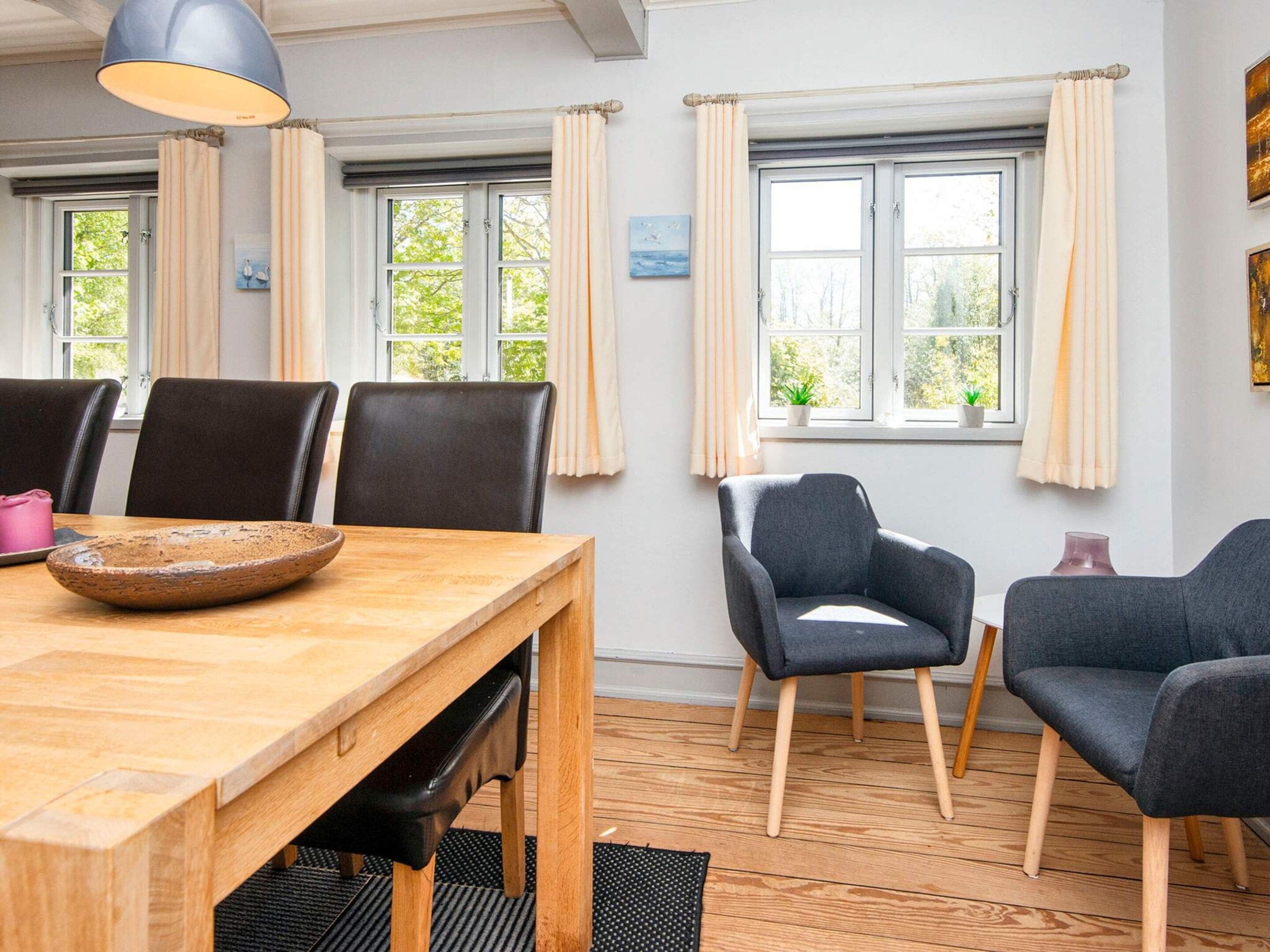 Un comedor con una mesa de madera, sillas negras y un cuenco. Hay ventanas con cortinas y vistas a zonas verdes.