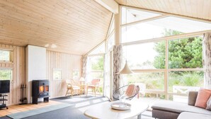 A cozy cabin interior with a fireplace, seating area, and large windows offering a view of greenery. - 4 Star Holiday Home in Henne (Henne)