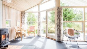 A wooden-paneled room with a wood stove, two wicker chairs, a black chair, and large windows. - 4 Star Holiday Home in Henne (Henne)