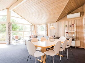 Une pièce avec un plafond en bois, une table ronde entourée de chaises, un canapé, une bibliothèque et de grandes fenêtres.