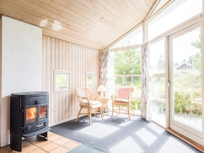 A room with a wood-burning stove, two wicker chairs, a table, and large windows.