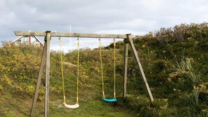 A wooden swing set in a grassy area with dense shrubbery and a house in the background. - 5 Person Holiday Home in Fano (Fano)