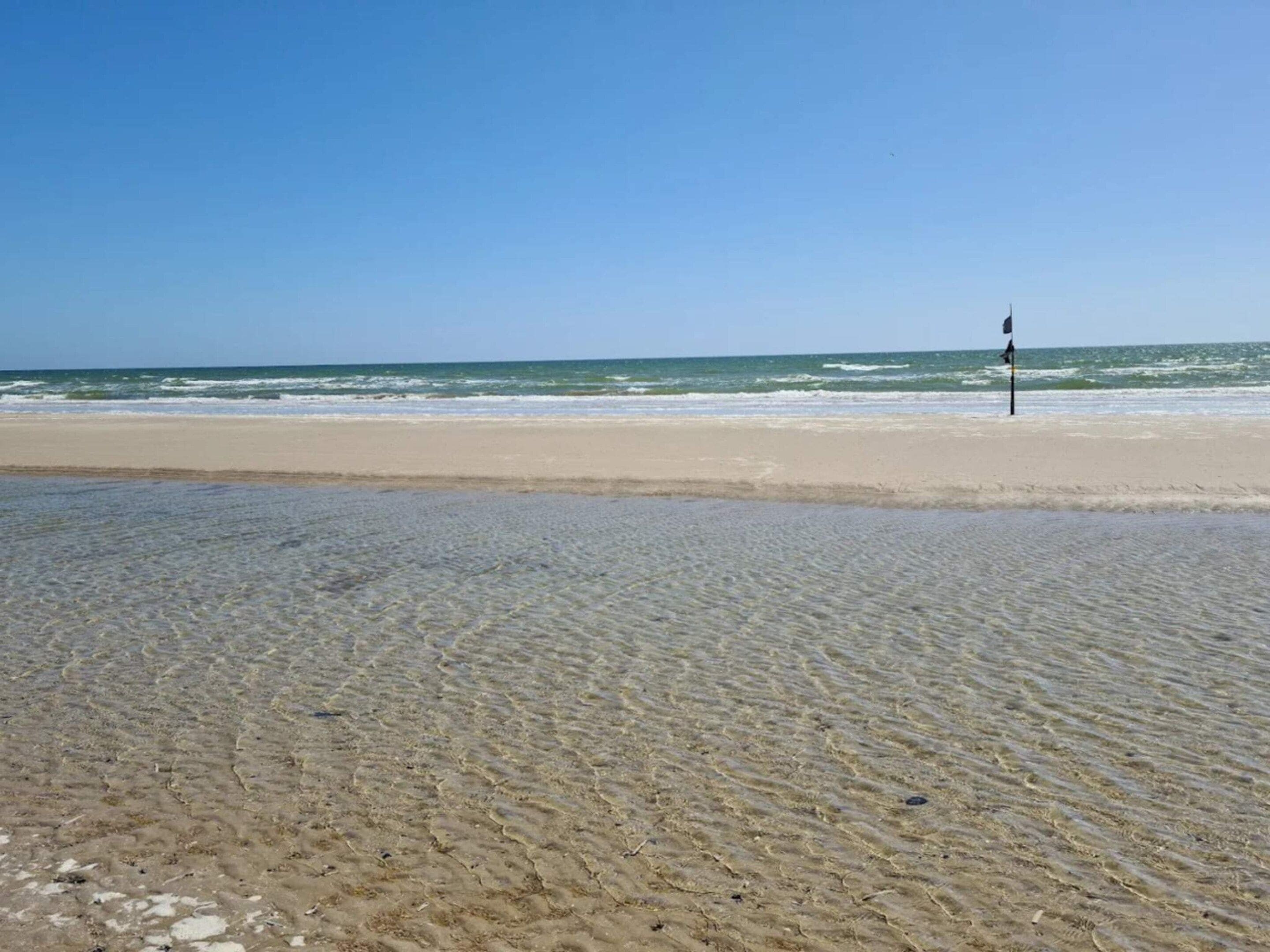 A beach with clear shallow waters and a flagpole in the distance.