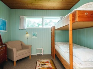 Ein Schlafraum mit Etagenbetten, Holzboden, einem Heizkörper, einem beigefarbenen Sessel und einem Fenster mit Vorhängen.