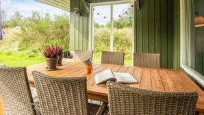 Une terrasse couverte avec une table en bois et des chaises en osier, une plante en pot et une vue sur la verdure environnante.