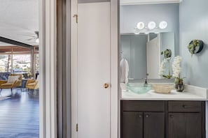 A bathroom with a glass bowl sink, a mirror with lights, and a vase with flowers.