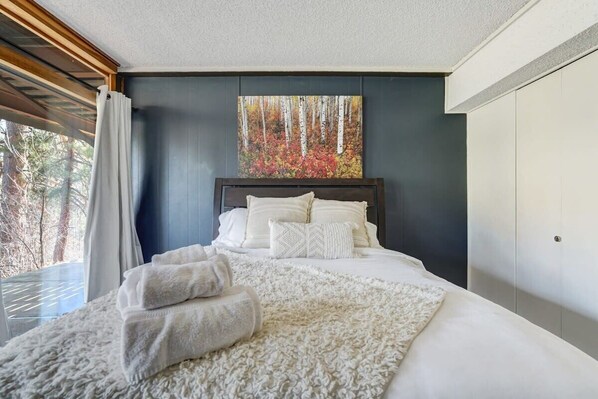 Une chambre à coucher moderne avec un grand lit, un tableau représentant une forêt et une vue sur les arbres par la fenêtre.