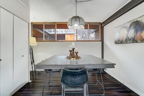 Un espace repas comprenant une longue table, deux chaises, un luminaire suspendu et une fenêtre donnant sur des arbres.