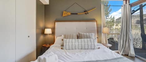 Une chambre à coucher avec un grand lit, une tête de lit, deux lampes de chevet, une fenêtre avec des rideaux et un objet décoratif fixé au mur.