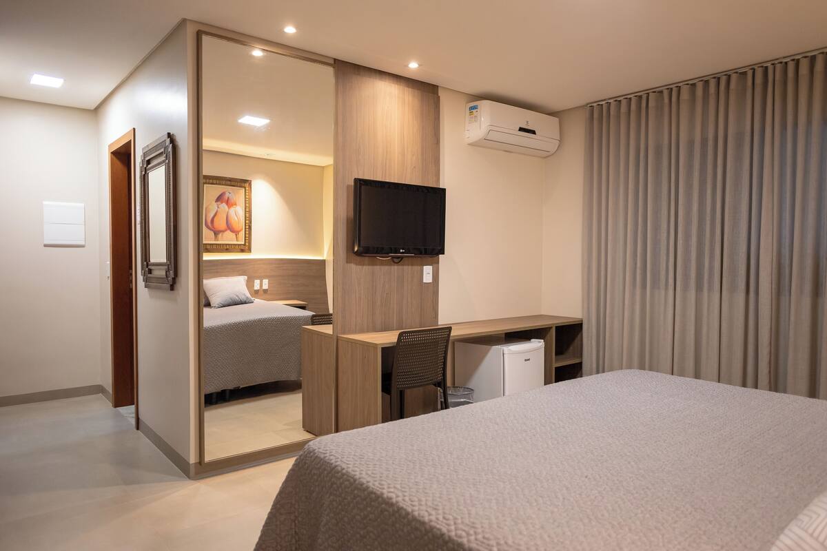 A hotel room with a bed, a desk, a chair, a television, and an air conditioning unit.