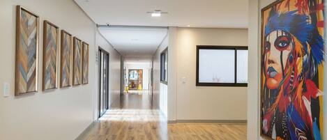 A hallway with wooden flooring, framed artworks on the walls, and a large colorful painting on the right.