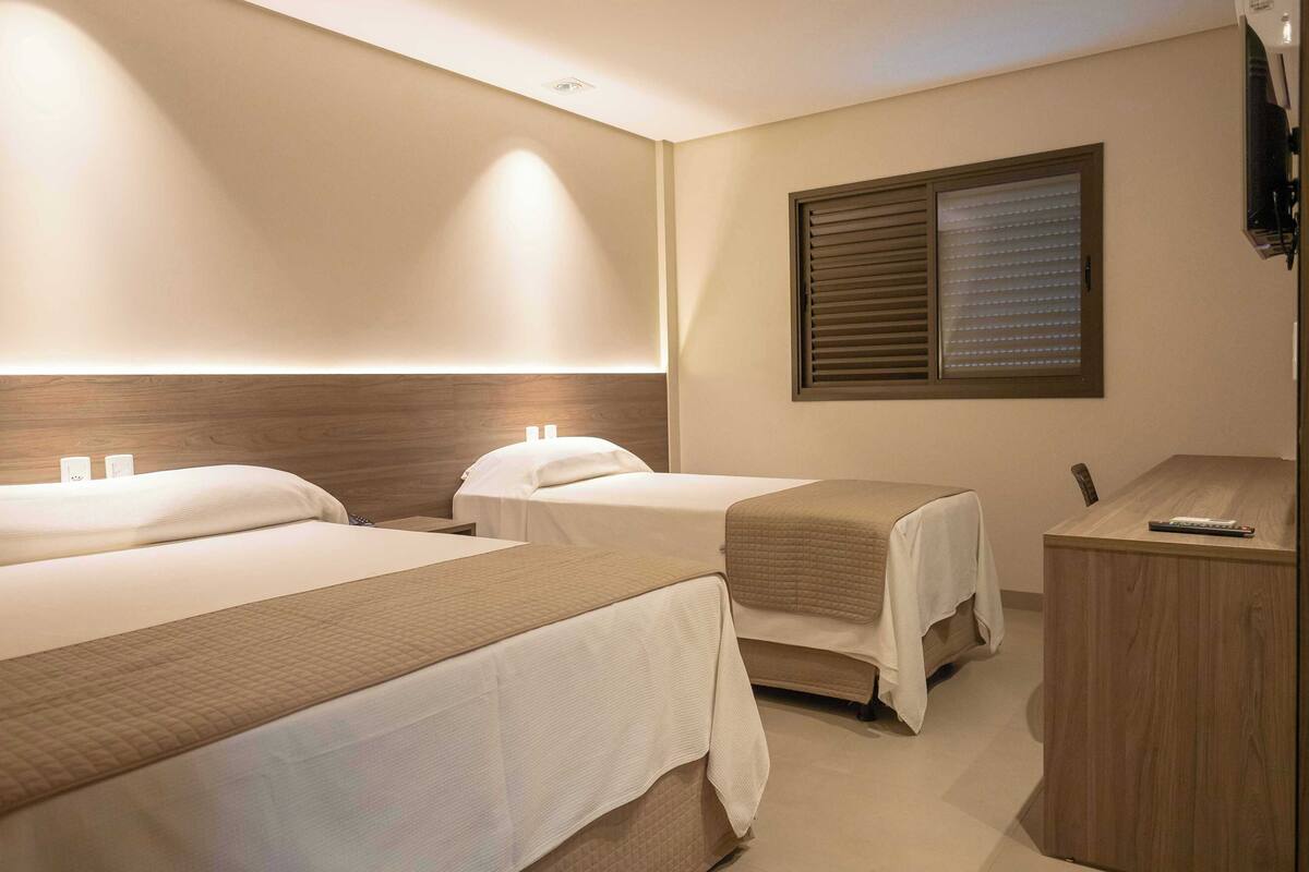 A hotel room with two beds, a desk, and a window with blinds.