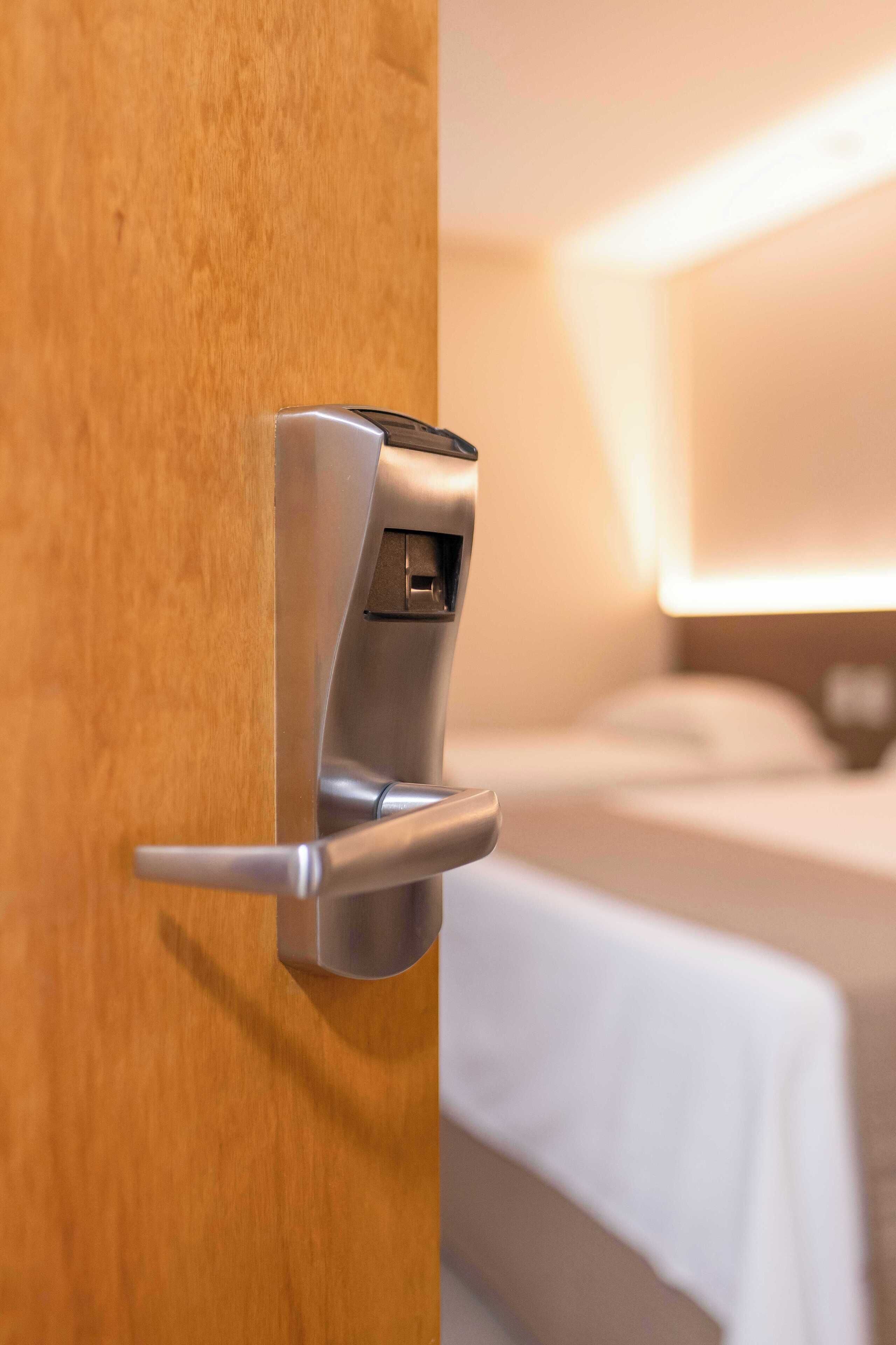 Une chambre d’hôtel moderne avec une porte en bois, un mini-bar intégré et un lit avec du linge de lit blanc.