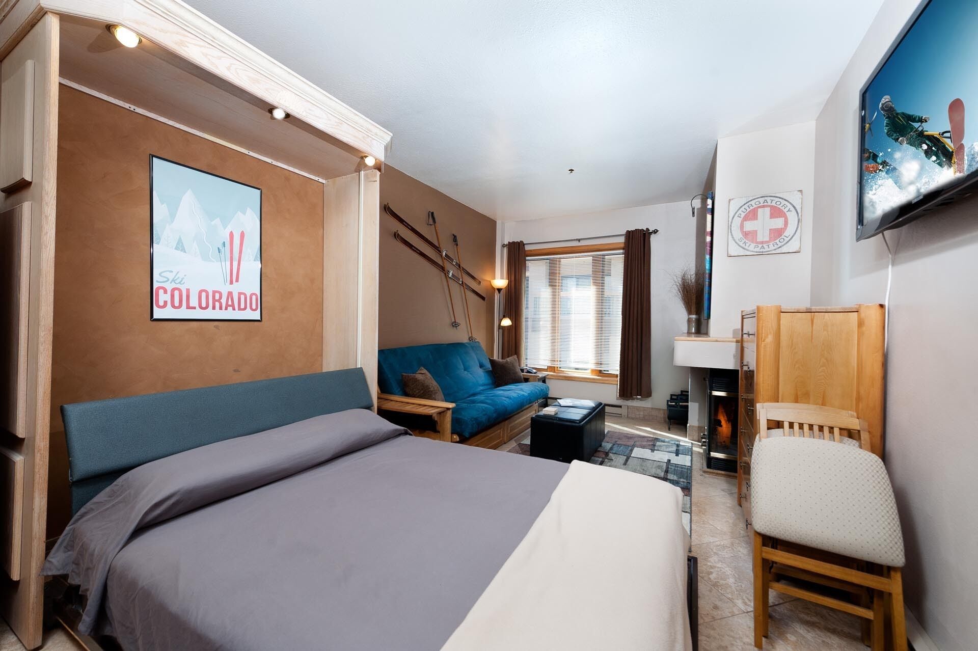 A cozy hotel room with a bed, a sofa, a chair, a fireplace, ski equipment, and a ski patrol sign.