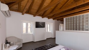 Een kamer met houten plafond, een flatscreen televisie aan de muur, een raam met uitzicht, een bank en een tafel met een notitieboekje en brillen.
