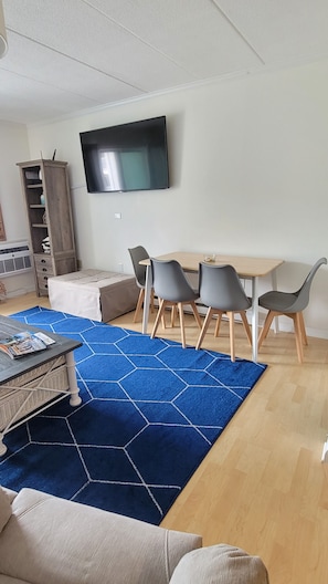 Uma sala de estar moderna com uma televisão de tela plana na parede, uma mesa de jantar de madeira com quatro cadeiras, um tapete hexagonal azul e um sofá.
