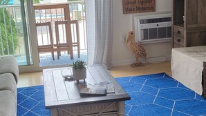 A living room with a blue patterned rug, a wooden coffee table with a plant, a large potted plant, a wooden cabinet, and a sign on the wall. - Sandy Jack's on 80th, 1 bedroom.  Steps to the beach and pool! Stocked! (Ocean City)