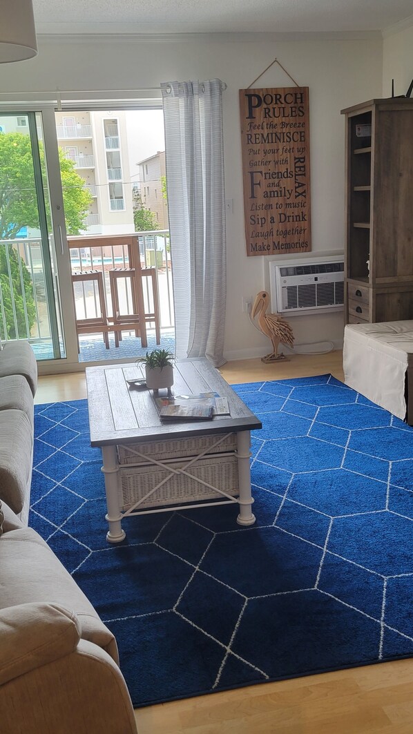 A living room with a blue patterned rug, a wooden coffee table with a plant, a large potted plant, a wooden cabinet, and a sign on the wall. - Sandy Jack's on 80th, 1 bedroom. Steps to the beach and pool! Stocked! (Ocean City)
