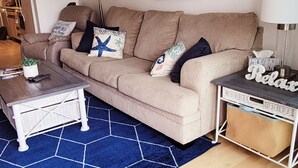 A living room with a beige sofa, a coffee table, a blue rug, and a painting of a bird on the wall. - Sandy Jack's on 80th, 1 bedroom.  Steps to the beach and pool! Stocked! (Ocean City)