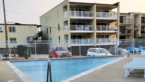 A swimming pool with a fenced area, a multi-story building with balconies, and a red vehicle parked nearby. - Sandy Jack's on 80th, 1 bedroom.  Steps to the beach and pool! Stocked! (Ocean City)