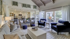 A spacious living room with a large sofa, a coffee table, and two armchairs. A kitchen is visible in the background with a dining area.