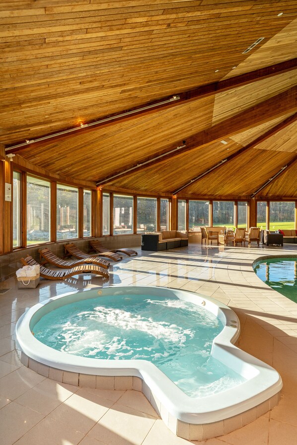 A spacious indoor pool area with a hot tub, wooden ceiling, and large windows offering views of the outdoors.