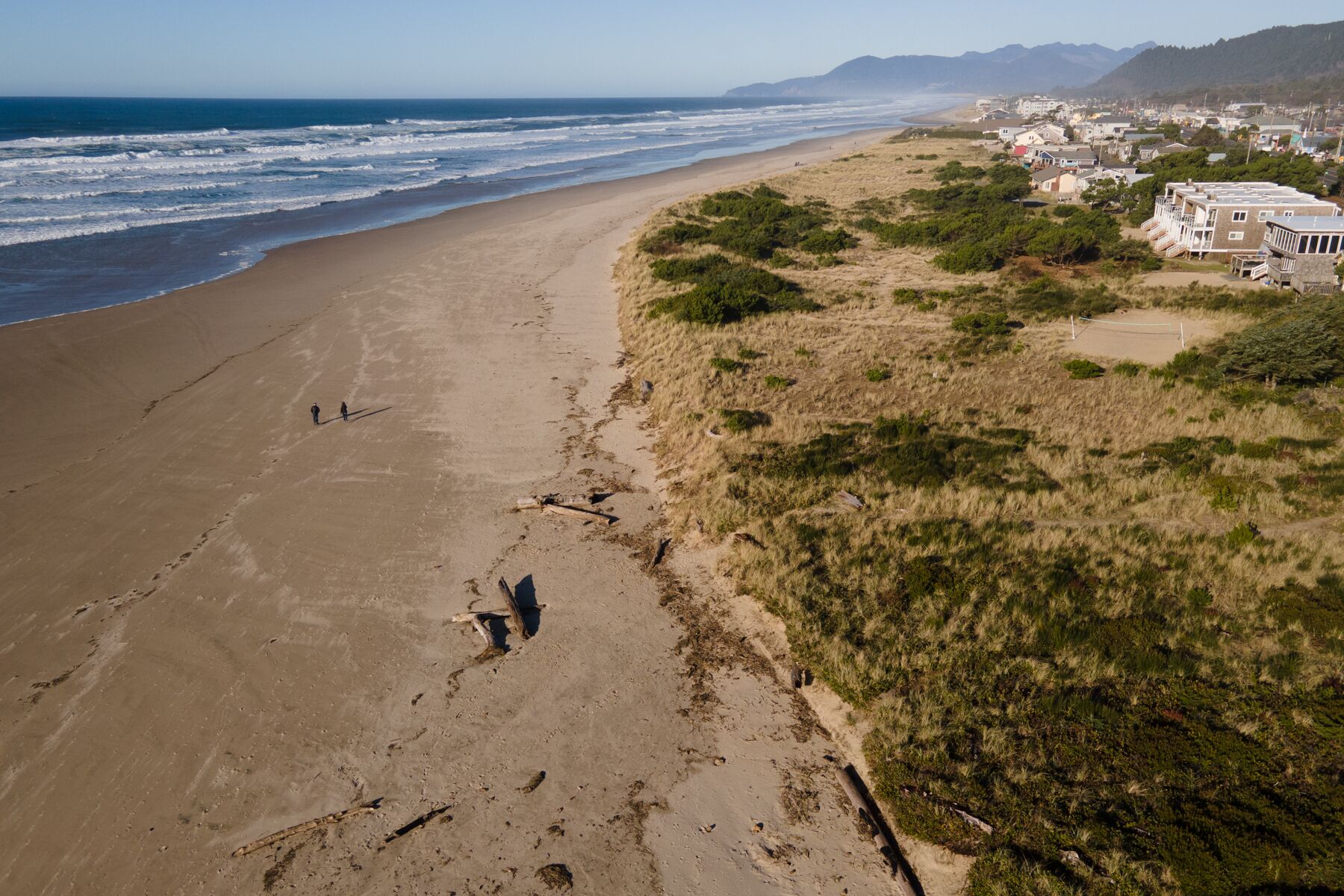 A coastal landscape with a sandy beach, a path leading to the shore, and a residential area with houses and greenery in the background.