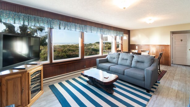 A living room with a large TV, a grey sofa, a wooden coffee table, and a striped rug. - Beachfront! Walk the short path to the Beach, or enjoy the waves from the Deck (Rockaway Beach)