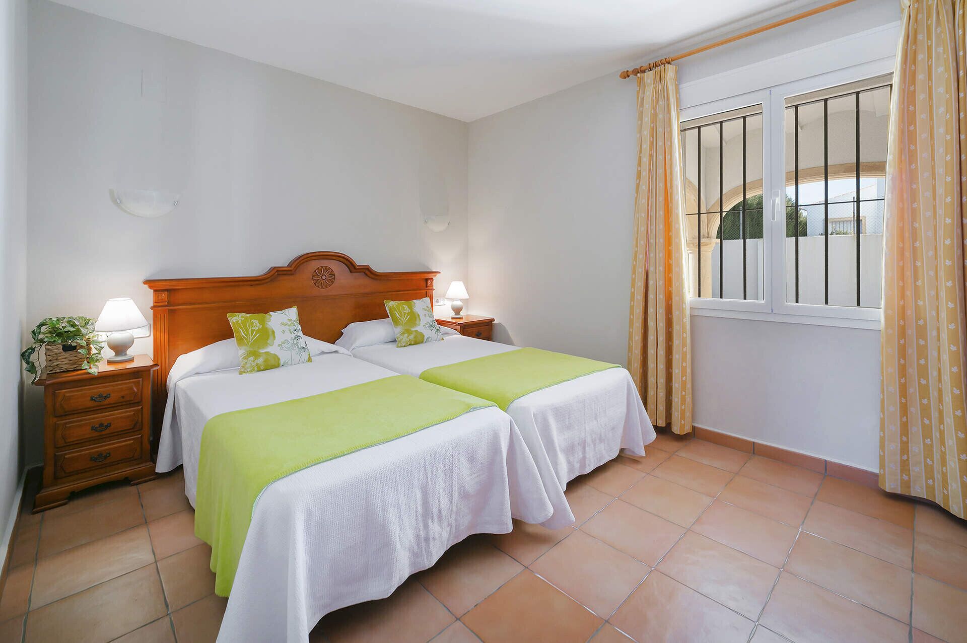 A bedroom with two beds, a wooden headboard, bedside tables with lamps, a window with curtains, and a tiled floor.