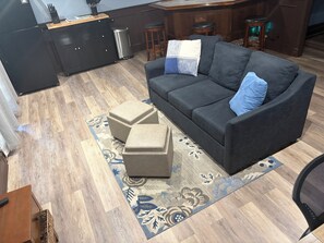 A modern living room with a dark grey sofa, two ottomans, a patterned rug, a wooden floor, a kitchen area with dark cabinetry, and a bar stool.