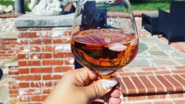 Uma mão segurando uma taça de vinho tinto com morango, com um pátio de tijolos e uma estátua ao fundo.