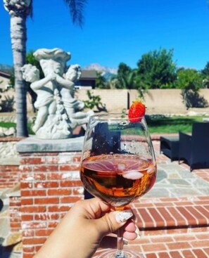 Una mano sostiene una copa de vino tinto con una fresa como adorno, frente a un patio de ladrillo y una estatua.