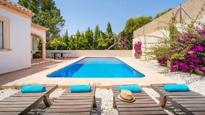 Une piscine entourée d’arbres et de plantes en fleurs, avec des chaises longues et une table à proximité.